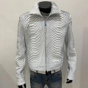 Pode incluir: Casaco branco com textura de pele de cobra, com fecho de correr e gola. O casaco é exibido num manequim, combinado com calças de ganga azuis e um cinto branco. O design do casaco apresenta um padrão escamoso.