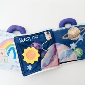 May include: A soft, open fabric book with a blue handle. One side features a rainbow, ABC blocks, and the text "Tiny Hands". The other side has a space theme with planets, stars, and the words "Blast Off".