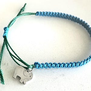 May include: Blue macrame bracelet with a silver elephant charm. The bracelet features a braided design in shades of blue and green, with an adjustable knot closure. The elephant charm is a simple, stylized silhouette.