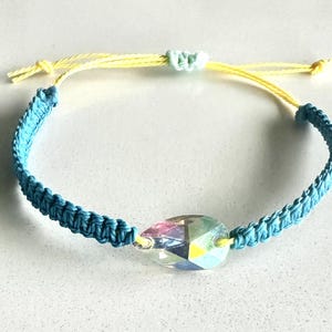 May include: A handmade bracelet featuring a teardrop-shaped iridescent crystal pendant. The bracelet has a blue macrame band and yellow adjustable cords. The crystal reflects multiple colors. Jewelry.