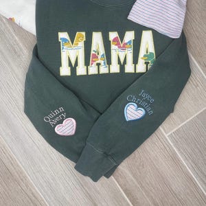May include: Dark green sweatshirt with the word "MAMA" embroidered in white with colorful duck designs. The sleeves feature embroidered names and heart designs. A striped sleeve is visible.