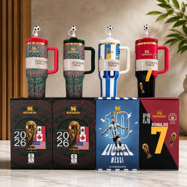 Pu&ograve; includere: Quattro bicchieri Stanley Quencher H2.0 con copripaglie a forma di pallone da calcio, ognuno con design e colori a tema. Sotto, quattro scatole di prodotto abbinate mostrano grafiche della Coppa del Mondo, immagini di giocatori e bandiere.