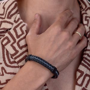 May include: Black and gray braided leather bracelet with a stainless steel clasp. The bracelet is worn on a wrist, and the person is wearing a patterned shirt with brown and beige colors.