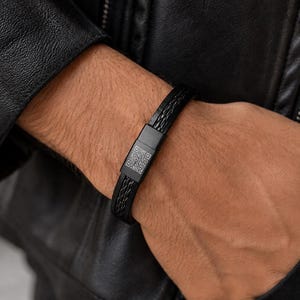 以下が含まれることがあります： 黒い編み込みレザーブレスレット、マットブラックの留め金付き。留め金には2つのQRコードが付いています。ブレスレットは手首に着用されており、背景には黒い革のジャケットがあります。