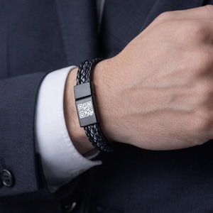Puede incluir: Pulsera de cuero trenzado negro con un cierre rectangular con un código QR. La pulsera se lleva en la muñeca, con una chaqueta de traje oscura y un puño de camisa blanca en el fondo.