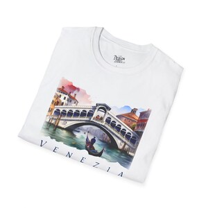 Könnte beinhalten: Weißes T-Shirt mit einem Aquarellgemälde der Rialtobrücke in Venedig, Italien, und einer Gondel. Das Wort "VENEZIA" ist unten in Dunkelblau gedruckt. Das Design Corner-Logo ist sichtbar.