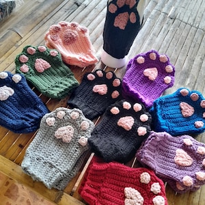 Puede incluir: Una colección de guantes de crochet hechos a mano con forma de pata en varios colores, incluyendo negro, azul, verde, rosa, gris, rojo y morado. Cada guante presenta un diseño de almohadilla de pata en un tono más claro, con un guante expuesto en un maniquí blanco.