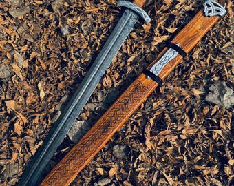 Könnte beinhalten: Ein Schwert und eine Schwertscheide werden gezeigt. Das Schwert hat eine silberne Klinge und einen Holzgriff. Die Schwertscheide ist aus Holz mit detaillierten Schnitzereien und silbernen Akzenten gefertigt. Schwert und Scheide liegen auf einem Bett aus braunen Blättern.