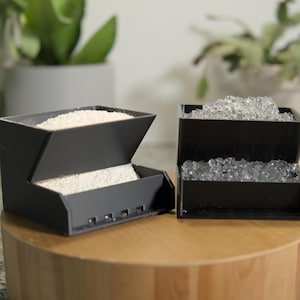 Puede incluir: Dos contenedores negros de diseño geométrico, uno lleno de gránulos blancos y el otro con cuentas transparentes. Están sobre un soporte de madera, con plantas al fondo. Los contenedores tienen un diseño angular.