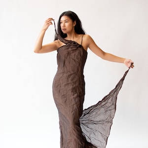 Op de afbeelding: Een vrouw draagt een lange, vloeiende bruine jurk met een ontwerp met één schouder. De jurk is gemaakt van een zachte, drapeerbare stof en heeft een vloeiende, etherische uitstraling.