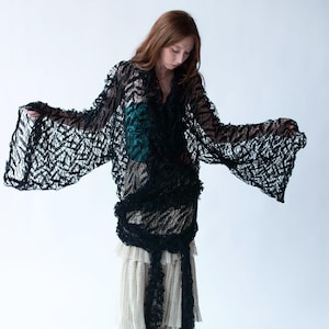 Può includere: Una donna indossa una blusa nera a maniche lunghe in pizzo sopra una gonna bianca a strati. La blusa è raccolta in vita e ha un aspetto fluido e drappeggiato.