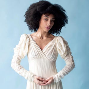 May include: A woman with curly black hair wearing a white, long-sleeved dress with a V-neckline and ruffled sleeves.