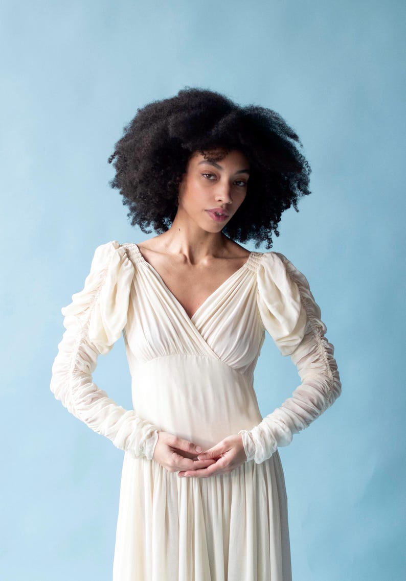 May include: A woman with curly black hair wearing a white, long-sleeved dress with a V-neckline and ruffled sleeves.