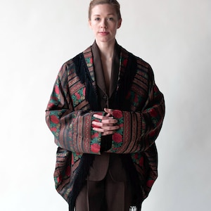 Può includere: Uno scialle fantasia con rose rosse e foglie verdi, frange nere e un completo marrone. Lo scialle è drappeggiato sulle spalle e sulle braccia. Lo sfondo è un muro bianco.