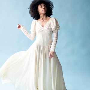 May include: A woman wearing a white, flowing dress with puffed sleeves. The dress has a V-neckline and a flowing skirt. The woman is standing on a light blue background.