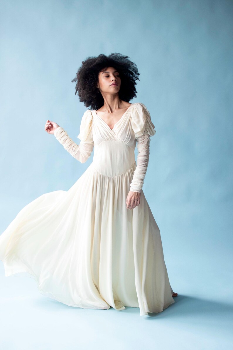 May include: A woman wearing a white, flowing dress with puffed sleeves. The dress has a V-neckline and a flowing skirt. The woman is standing on a light blue background.