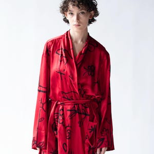 Puede incluir: Una bata roja con un estampado floral negro. La bata tiene mangas largas y un cintur&oacute;n a juego. La tela parece ser sedosa. La bata la lleva una persona con el pelo corto y rizado.