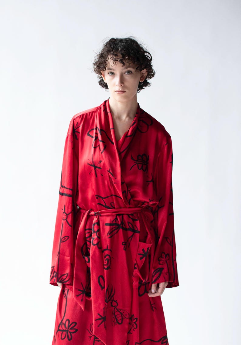 Puede incluir: Una bata roja con un estampado floral negro. La bata tiene mangas largas y un cintur&oacute;n a juego. La tela parece ser sedosa. La bata la lleva una persona con el pelo corto y rizado.