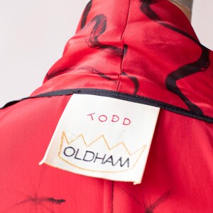Puede incluir: Primer plano de una prenda roja con trazos abstractos negros. La prenda tiene un cuello alto y una etiqueta que dice "TODD OLDHAM" en letras rojas y negras. La etiqueta es de color crema con un dise&ntilde;o de corona dorada.