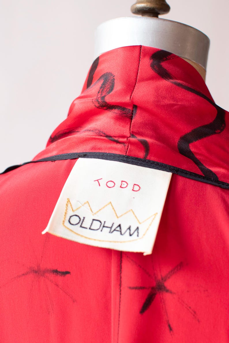Puede incluir: Primer plano de una prenda roja con trazos abstractos negros. La prenda tiene un cuello alto y una etiqueta que dice "TODD OLDHAM" en letras rojas y negras. La etiqueta es de color crema con un dise&ntilde;o de corona dorada.