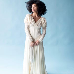 May include: A woman wearing a long, white, flowing dress with puffed sleeves. The dress has a V-neckline and a fitted bodice. The woman is standing on a light blue background.