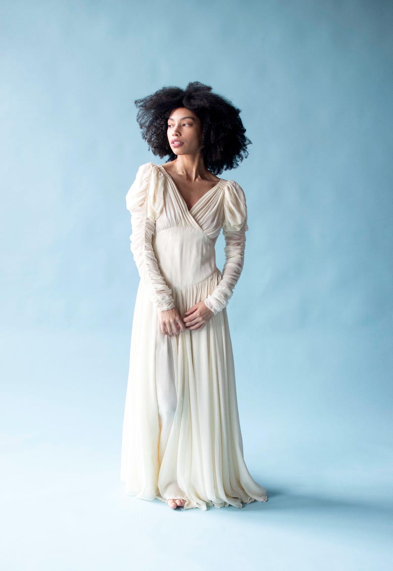 May include: A woman wearing a long, white, flowing dress with puffed sleeves. The dress has a V-neckline and a fitted bodice. The woman is standing on a light blue background.