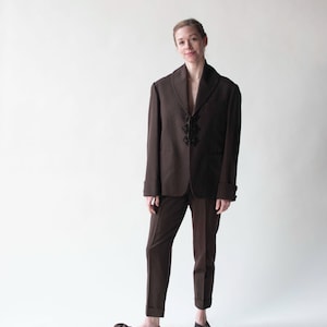 Può includere: Un abito marrone con collo a scialle e bottoni decorativi. L'abito è indossato da una persona con capelli castano chiaro e sta in piedi su uno sfondo bianco.