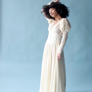 May include: A woman wearing a white, long-sleeved gown with a flowing skirt and puffed sleeves. She is standing against a light blue background.