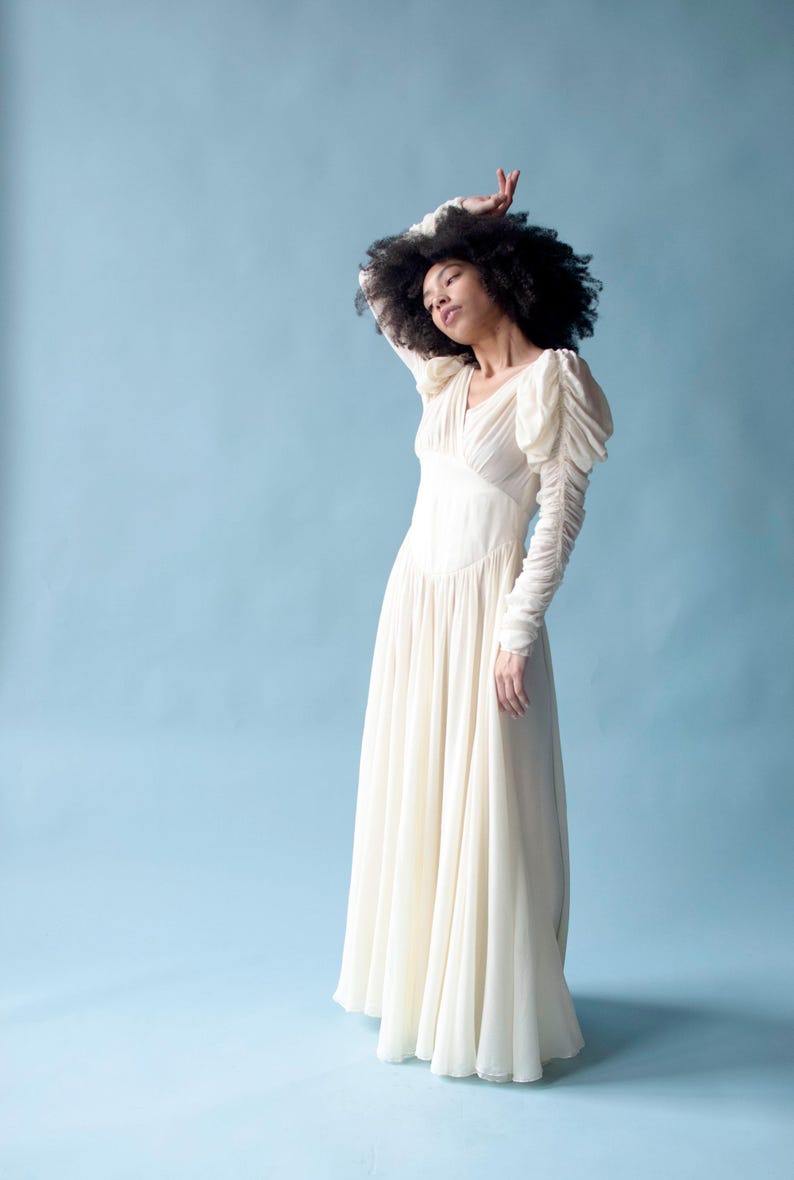 May include: A woman wearing a white, long-sleeved gown with a flowing skirt and puffed sleeves. She is standing against a light blue background.