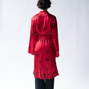 Puede incluir: Una bata roja vibrante con dise&ntilde;os florales y abstractos en negro. La bata tiene mangas largas y un cintur&oacute;n a juego. La persona lleva pantalones y zapatos negros. El fondo es blanco liso.