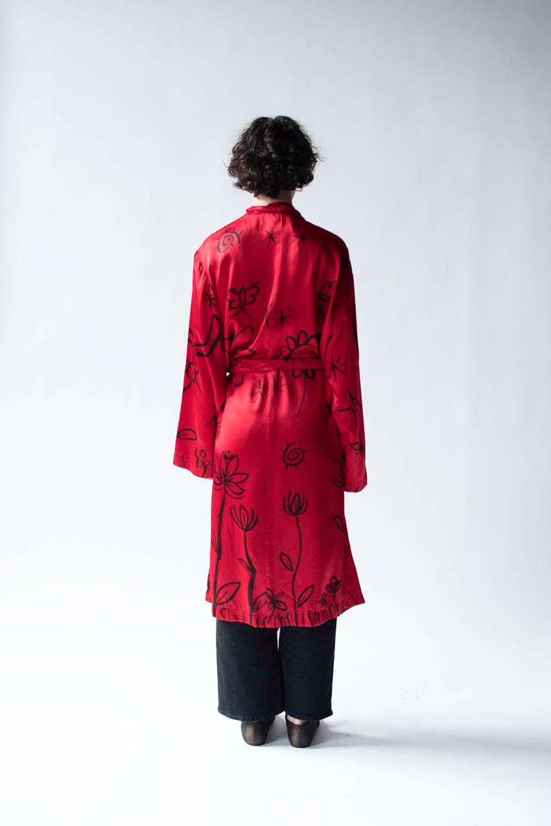 Puede incluir: Una bata roja vibrante con dise&ntilde;os florales y abstractos en negro. La bata tiene mangas largas y un cintur&oacute;n a juego. La persona lleva pantalones y zapatos negros. El fondo es blanco liso.