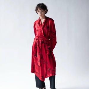 Puede incluir: Un vestido envolvente rojo con estampados florales negros. El vestido tiene cintur&oacute;n y mangas largas. La persona lleva pantalones negros y zapatos marrones. El fondo es blanco.