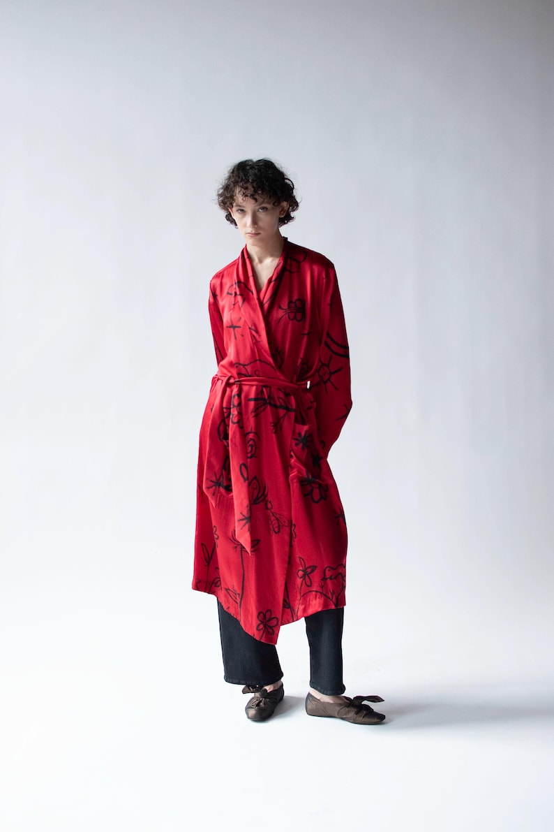 Puede incluir: Un vestido envolvente rojo con estampados florales negros. El vestido tiene cintur&oacute;n y mangas largas. La persona lleva pantalones negros y zapatos marrones. El fondo es blanco.