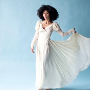 May include: A woman with curly dark hair wearing a long, flowing white dress with puffed sleeves. The dress has a V-neckline and a flowing skirt. The woman is standing on a light blue background.