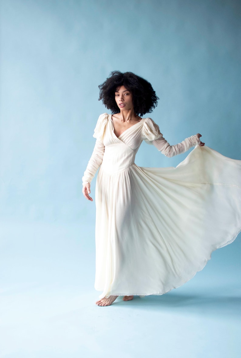 May include: A woman with curly dark hair wearing a long, flowing white dress with puffed sleeves. The dress has a V-neckline and a flowing skirt. The woman is standing on a light blue background.