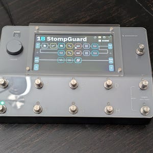 May include: A gray electronic device with the text "1B StompGuard" on a screen. The device has multiple silver buttons, a volume knob, and a screen displaying a graphic interface. The device is on a dark wooden surface.