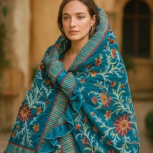 Könnte beinhalten: Ein türkisfarbener Samtumhang mit floralen Stickereien in Orange, Rot und Gelb. Der Umhang hat einen gerüschten Rand und einen gestreiften Besatz in Rot und Gold. Ein luxuriöses Accessoire.