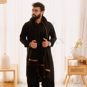 May include: A person wearing a black outfit, including a long-sleeved shirt and loose pants. A black scarf with brown stripes is draped over the shoulders. The outfit is paired with black shoes. The background includes a woven lamp and a wooden side table.
