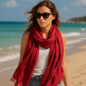 May include: A woman on a beach wearing a vibrant red scarf with fringed edges. The scarf is wrapped around the neck and shoulders. She is wearing sunglasses. The background shows the sea and a clear sky.