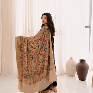 Puede incluir: Un chal beige con intrincados bordados que representan pájaros coloridos y motivos florales. El chal tiene un borde ancho y detallado. El fondo es de color neutro.