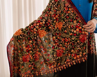 Puede incluir: Chal negro con intrincados bordados florales en rojo, naranja y verde. El chal tiene un borde negro y un patrón detallado. El diseño se establece sobre un fondo negro, creando un contraste llamativo. El chal está drapeado sobre los hombros.