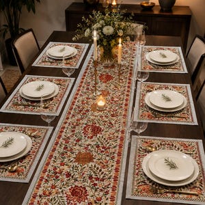 May include: A dining table set featuring a long rectangular table runner and six matching placemats, all decorated with a detailed floral pattern in shades of red, orange, and green on a cream background, arranged with white plates and glassware.