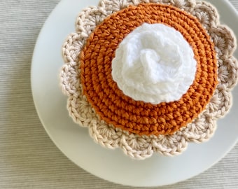 May include: A crocheted pumpkin pie with a white whipped cream topping. The pie is on a white plate with a brown background.