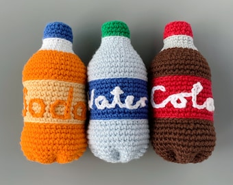 Op de afbeelding: Drie gehaakte flessen, een oranje, een blauwe en een bruine, met de woorden "Soda", "Water" en "Cola" erop.