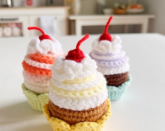 May include: Three crocheted cupcakes with red cherries on top. The cupcakes are layered in different colors, including orange, purple, and brown. The cupcakes are sitting on a white surface.
