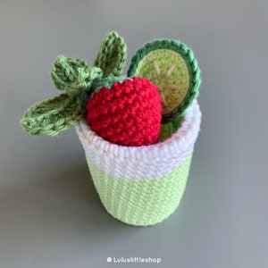 May include: A crocheted green cup with a white rim filled with a red crocheted strawberry and a green crocheted cucumber slice.