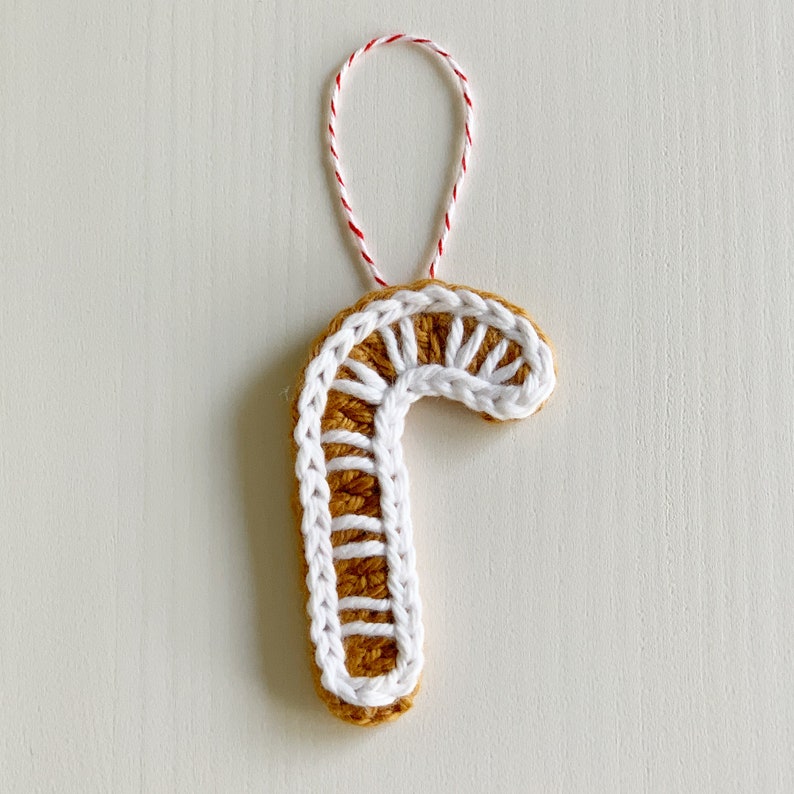 K&ouml;nnte beinhalten: Ein geh&auml;kelter Zuckerstangen-Anh&auml;nger mit einem wei&szlig;en und braunen Design. Der Anh&auml;nger h&auml;ngt an einer rot-wei&szlig;en Schnur.