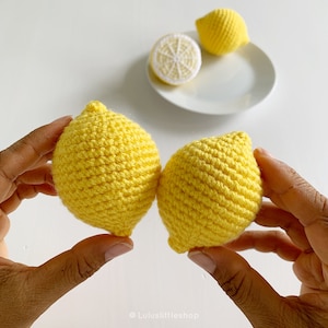 May include: Two crocheted yellow lemons held in hands. The lemons are made with a textured stitch and have a realistic shape.