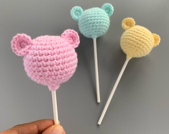 Op de afbeelding: Drie gehaakte lolly's in de vorm van berenhoofdjes in roze, blauw en geel. De lolly's zijn op witte stokjes.