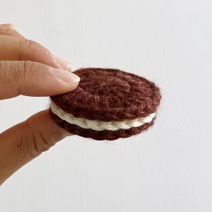 May include: A crocheted chocolate sandwich cookie with white frosting. The cookie is brown and has a textured surface.